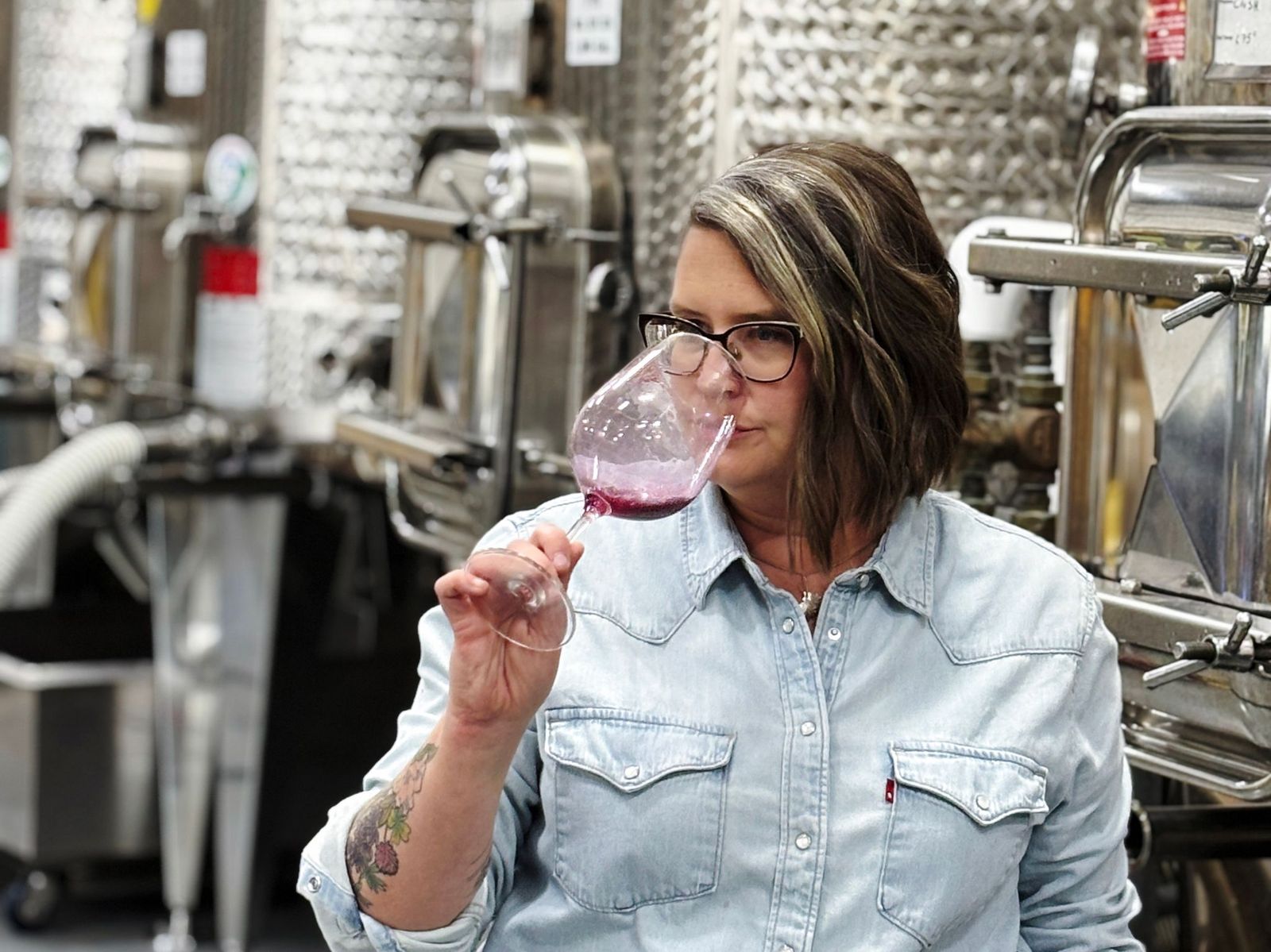 A portrait of Quattro Theory Wines winemaker, Emily Hanes, in the cellar in front of stainless steel tanks, sampling some wine from a wineglass